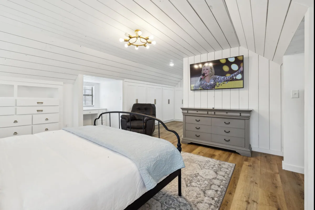 A serene guest bedroom with a bed dressed in light blue bedding, a gray dresser, a wall-mounted television, and a comfortable reclining chair. The room is illuminated by soft, natural light.