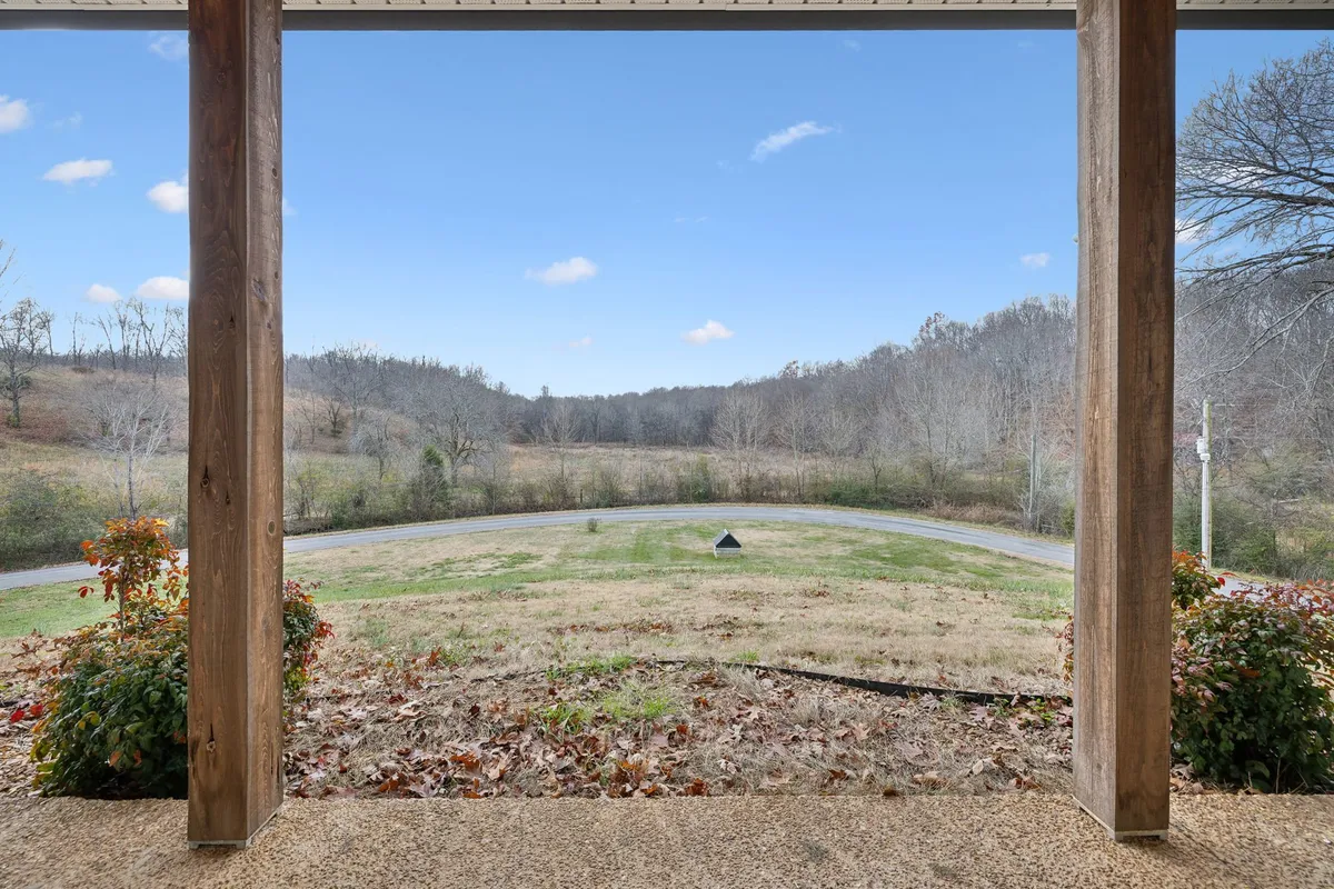 An expansive landscape stretches out from the vantage point of the front porch, with porch posts framing the view, a curving driveway, and gentle hills in the background. The scene is serene, ideal for sipping morning coffee, watching sunsets, or planning nature walks.