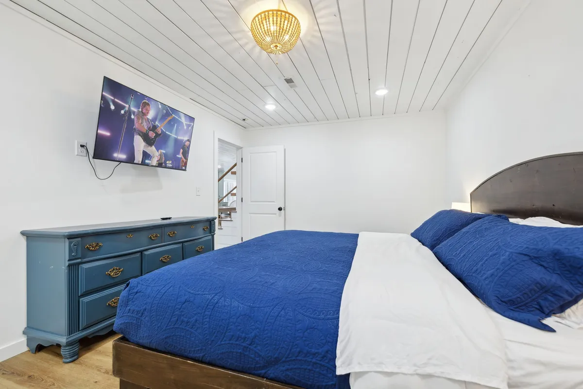 A spacious bedroom featuring a king-sized bed with a deep blue quilted bedspread, a wooden dresser, a large flat-screen Smart TV, and a modern chandelier. The room is flooded with natural light and exudes modern comfort.