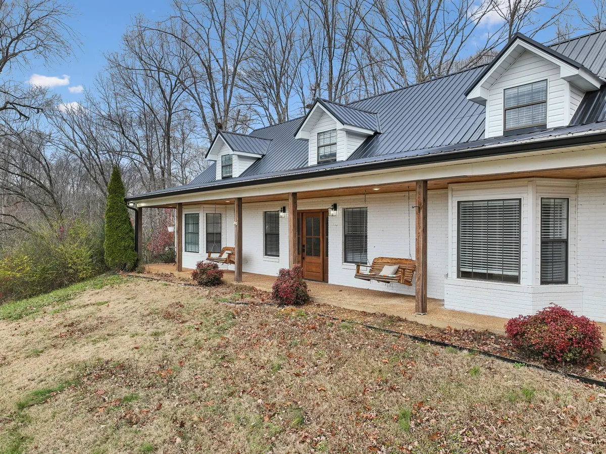The image showcases the welcoming front porch of a modern cabin-style home, framed by wooden benches perfect for relaxing. The lush, landscaped yard leads up to the inviting front door, all set beneath a serene, open sky—hinting at sunset views and a tranquil outdoor setting.