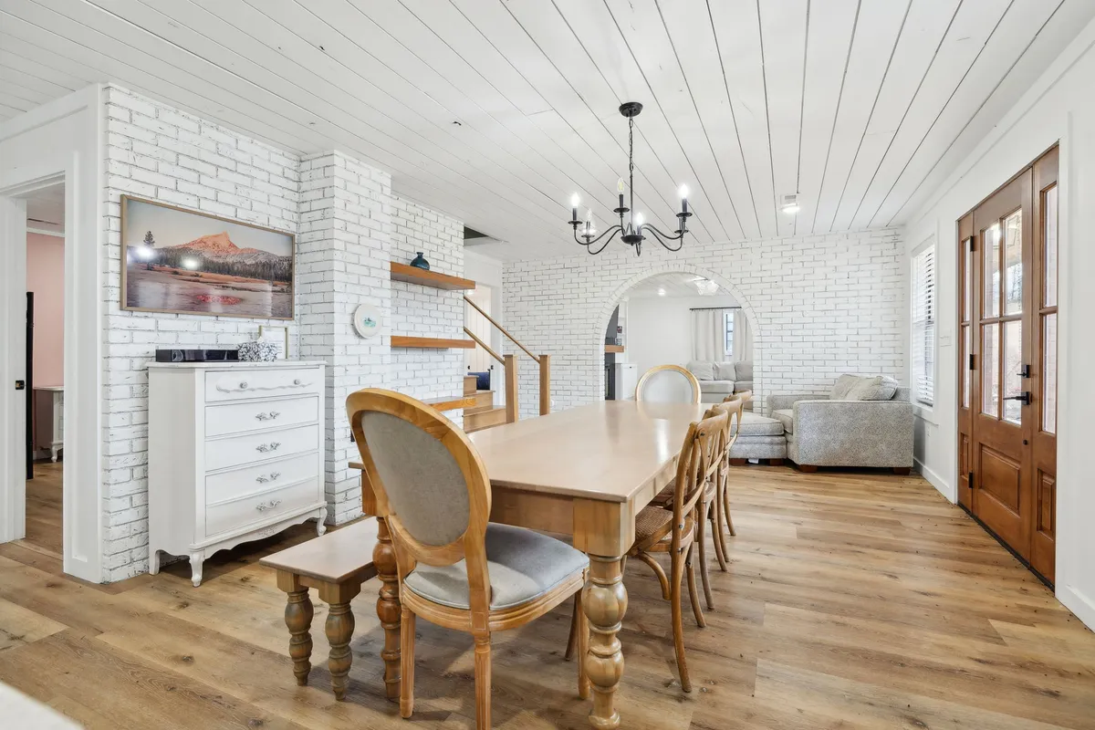 An interior view of the spacious dining table, complemented by a warm chandelier, a feature brick wall, and large windows that bring in natural light. The setting is ideal for sharing meals, whether cooking in, enjoying takeout, or celebrating with family and friends.