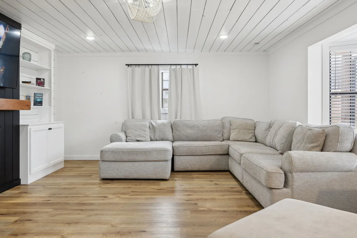 A cozy living area featuring a large sectional sofa, a sparkling chandelier overhead, and a wall-length bookshelf. The room feels warm and accommodating, perfect for group relaxation, storytelling, or a game night after a day spent outdoors.