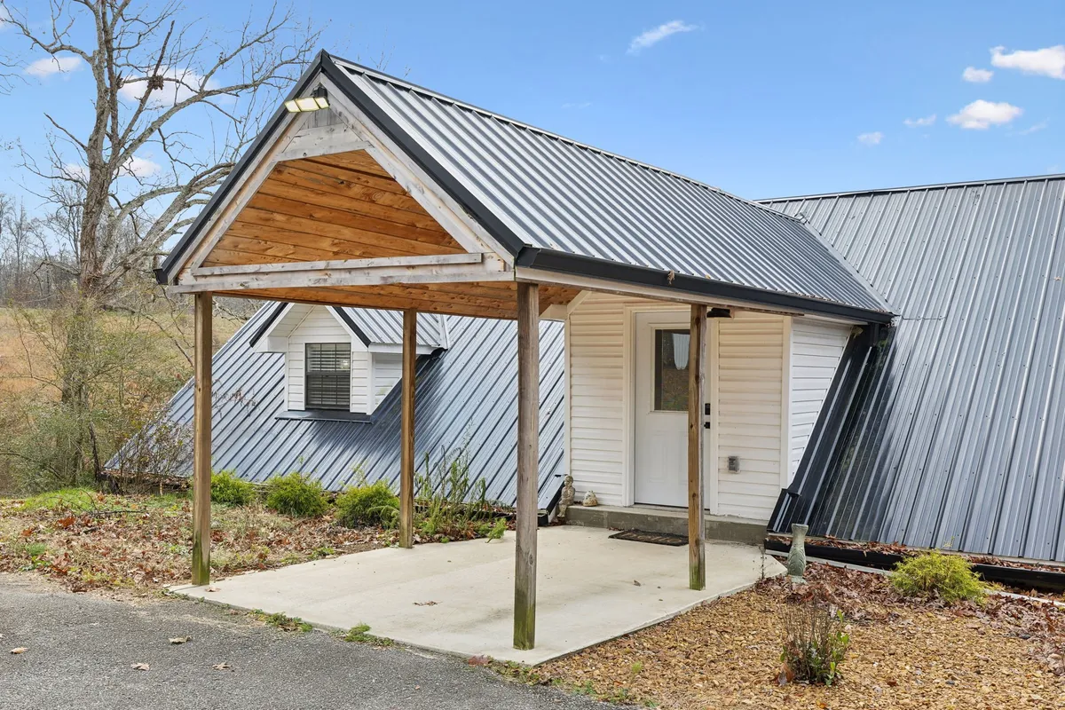 This image showcases the home's covered entrance with a metal roof, wooden beams, and attractive landscaping. The driveway is prominently visible, providing a welcoming approach for guests and demonstrating the ample parking available for groups.